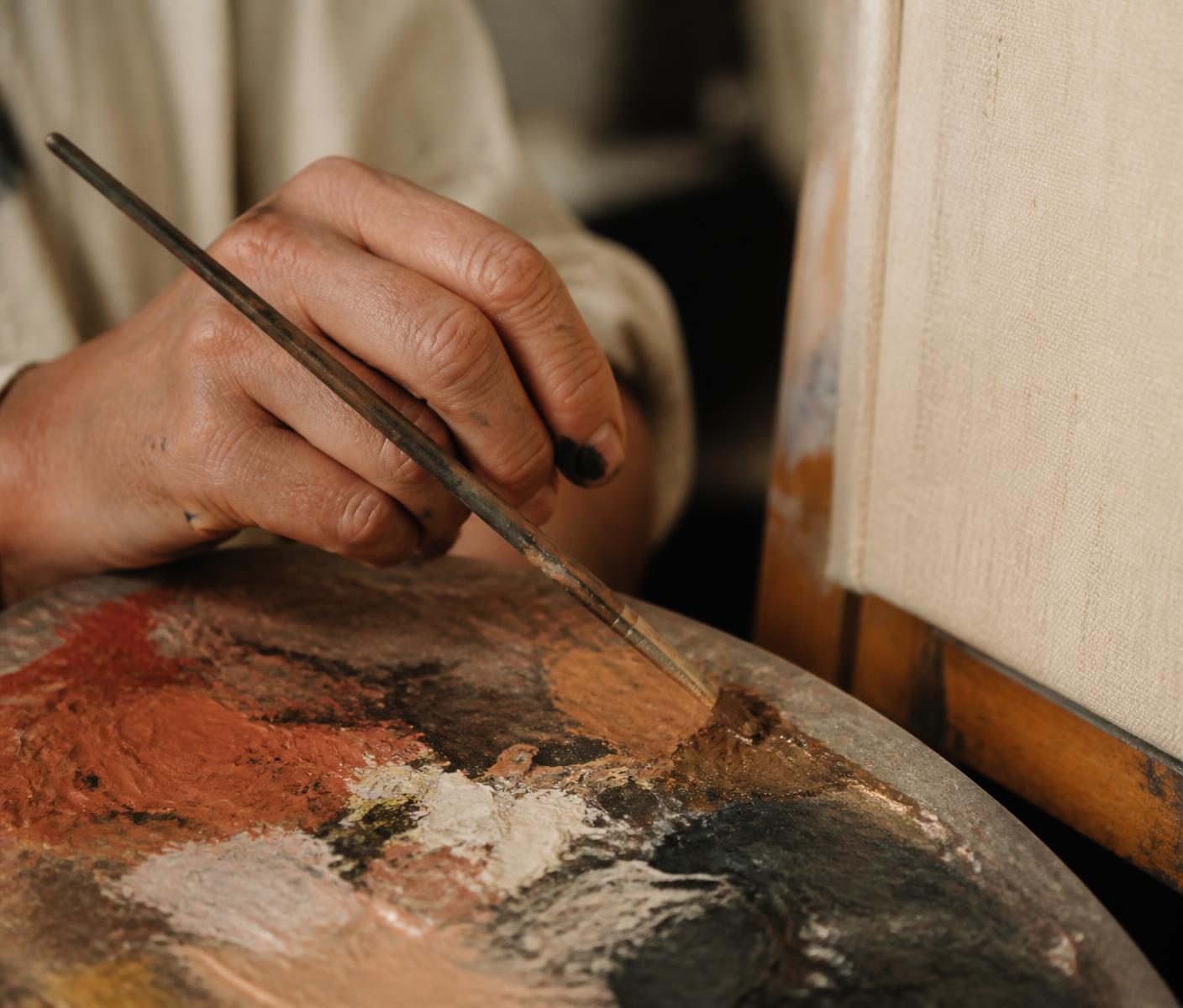 An artist's hand holding a brush above a palette of terracotta, bronze and charcoal paint